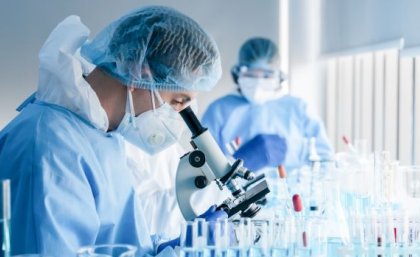 The researchers found Australia is falling behind international benchmarks for funding of basic research. Image: Adobe. A scientist in cap, gown and mask stands in profile looking down through a microscope as a second scientist looks on in the background. They are in a lab.